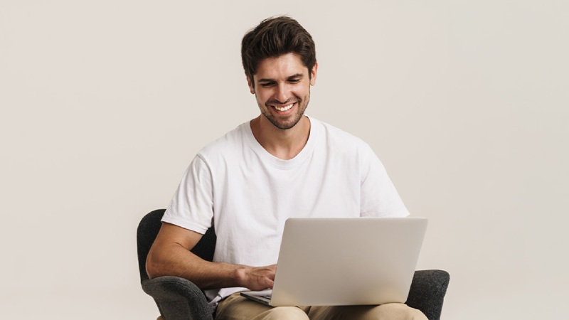 Smiling man in a white T-shirt using a laptop while sitting in a chair