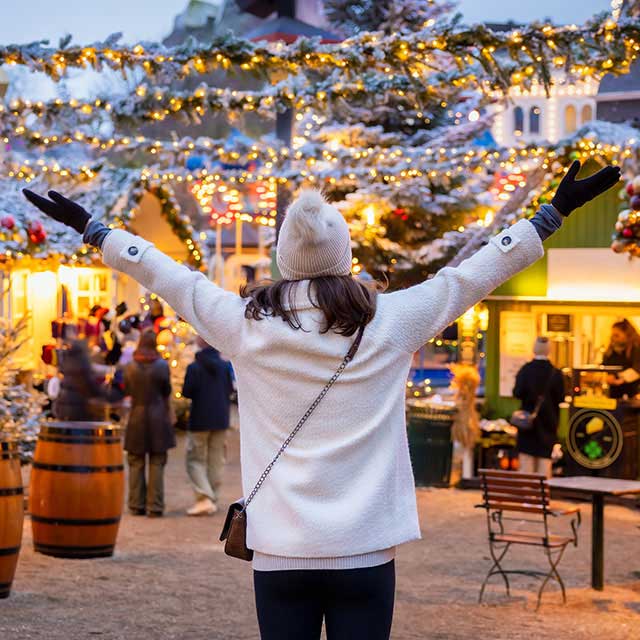 A woman in a white coat and beanie joyfully raises her arms at a festive Christmas market, adorned with glowing lights and decorated trees.