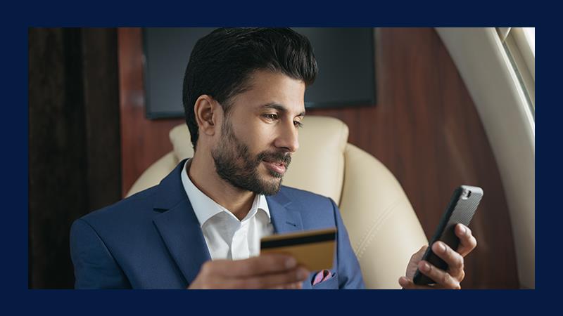 A man in a blue suit sits in an airplane, holding a credit card and a smartphone. He appears focused, suggesting online shopping or payment.