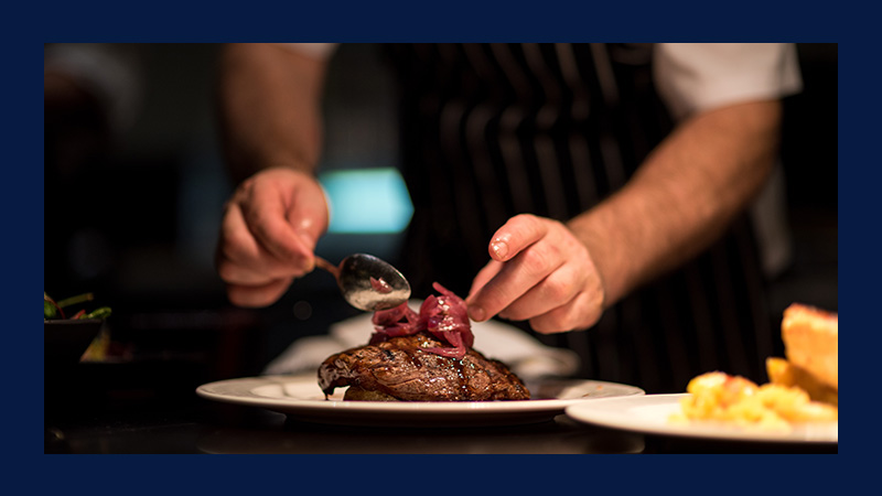 Chef garnishing steak with caramelized onions using a spoon. The scene is intimate and professional, highlighting culinary expertise and gourmet cooking.
